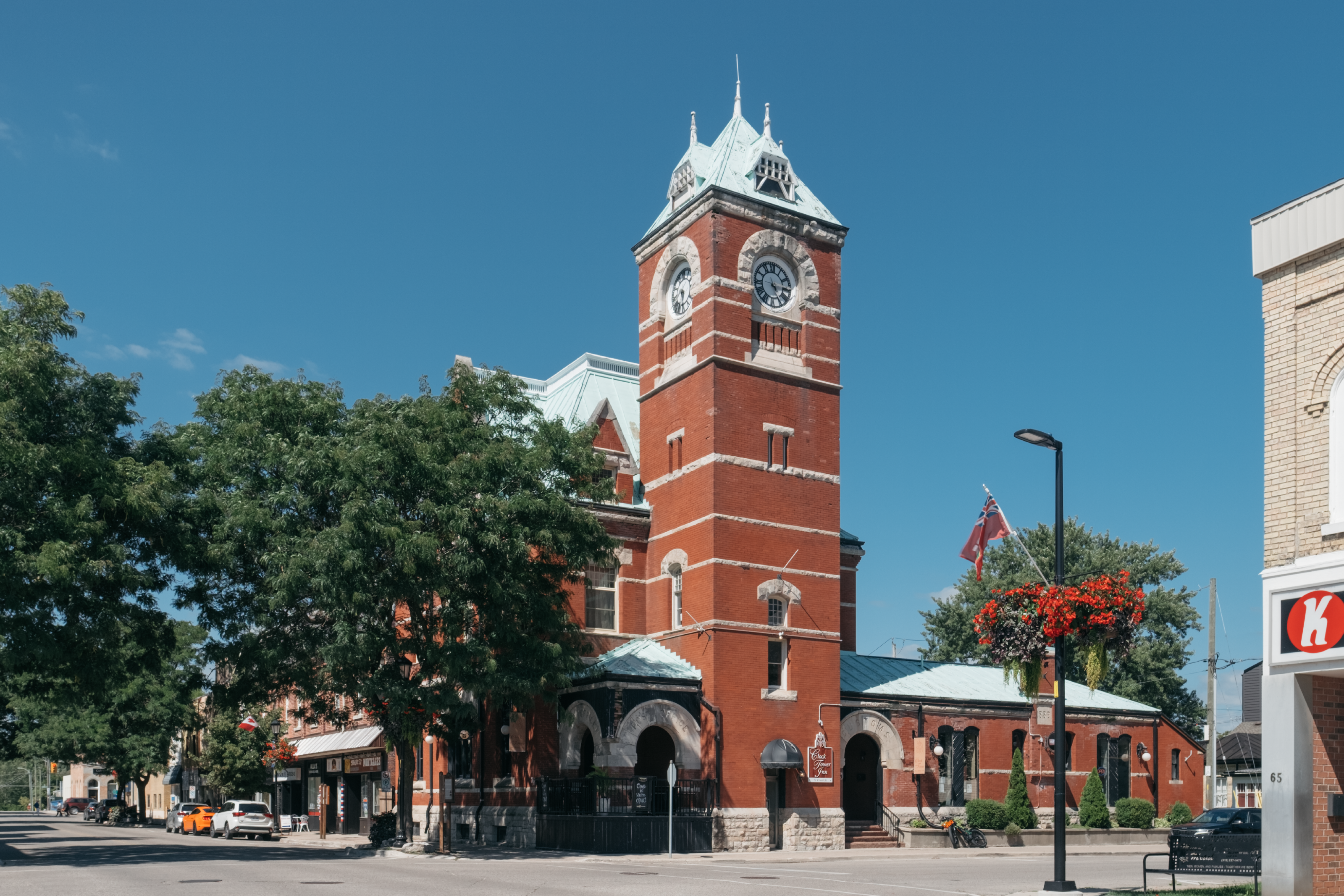 Tall, red brick clock tower on the corner of an intersection, adjoined by a one-storey brick building on the right and a three-storey brick building on the left. 